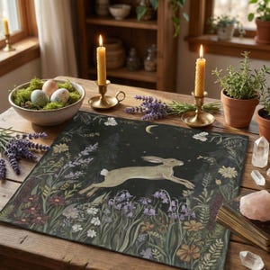 May include: A decorative placemat featuring a running hare illustration in a floral and forest setting, with a crescent moon in the night sky. The placemat is on a wooden table, with candles, eggs, and plants.