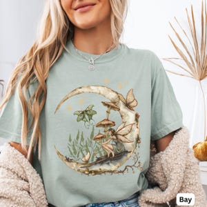 May include: A sage green t-shirt featuring a graphic print of a crescent moon, a fairy with wings, mushrooms, butterflies, and tall grass. The design includes small star shapes and delicate branch details. The shirt has a crew neckline.