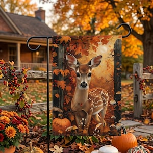 May include: A rectangular garden flag featuring a fawn standing in an autumn forest. The scene includes orange and brown fall foliage, a wooden fence, a pumpkin, and small mushrooms. The flag is suspended from a black metal garden stake.