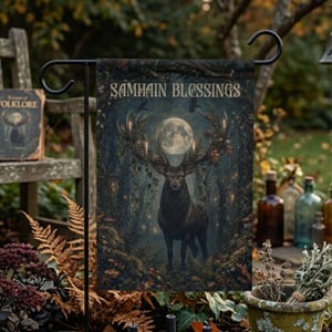 May include: A rectangular garden flag featuring a dark forest scene with a large stag with expansive antlers centered beneath a full moon. The text SAMHAIN BLESSINGS is printed in a light, serif font at the top of the flag, which hangs on a black metal stand.