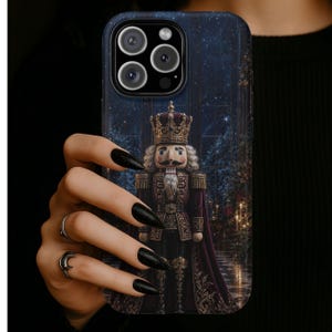 May include: A smartphone case featuring a detailed illustration of a nutcracker king wearing a crown and a deep burgundy cape. The background depicts a dark, starry night scene with a festive atmosphere, including subtle hints of holiday decor.
