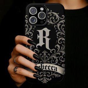 May include: Black phone case with an ornate, swirling design and the letter "R" in the center. The name "Rebecca" is displayed on a banner at the bottom. The case has a glossy finish.