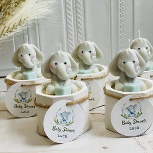 May include: Set of six white ceramic candle holders with elephant-shaped candles. Each holder has a round tag with a blue and green elephant illustration and the text "Baby Shower Luca".