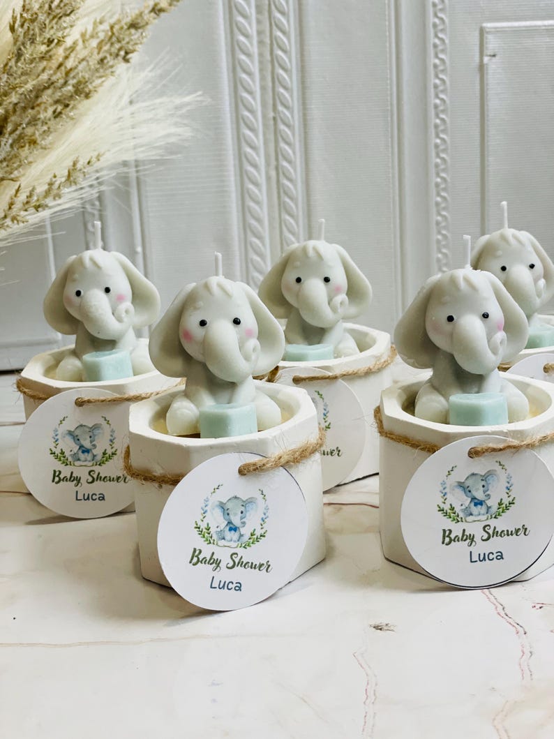 May include: Set of six white ceramic candle holders with elephant-shaped candles. Each holder has a round tag with a blue and green elephant illustration and the text "Baby Shower Luca".