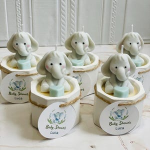 May include: Six small white candles shaped like elephants holding a blue heart. Each candle is in a white container with a brown twine bow and a round white tag with a blue elephant design and the text "Baby Shower Luca".