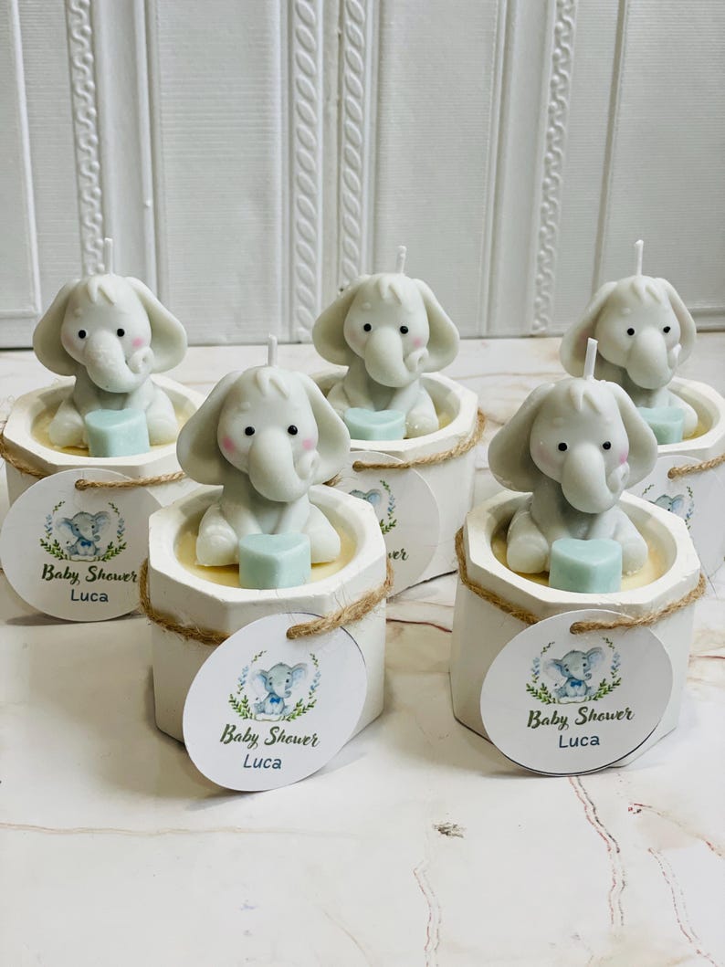 May include: Six small white candles shaped like elephants holding a blue heart. Each candle is in a white container with a brown twine bow and a round white tag with a blue elephant design and the text "Baby Shower Luca".