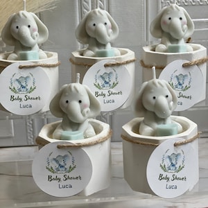 May include: Five white favor boxes with a small elephant candle inside. Each box has a round tag with a blue and green elephant design and the text "Baby Shower Luca".
