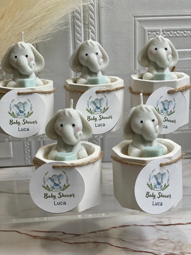 May include: Five white favor boxes with a small elephant candle inside. Each box has a round tag with a blue and green elephant design and the text "Baby Shower Luca".