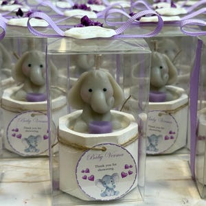 May include: A set of small, clear boxes with purple ribbon bows, each containing a white ceramic elephant candle with a purple base. The boxes have a label that says "Baby Verma" and "Thank you for showering".