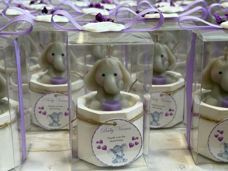 May include: A set of small, clear boxes with purple ribbon bows, each containing a white ceramic elephant candle with a purple base. The boxes have a label that says "Baby Verma" and "Thank you for showering".