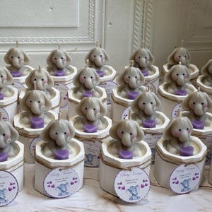 May include: A collection of small, white, hexagonal boxes with a gray elephant candle inside each. The boxes have a brown twine bow and a small tag that says "Baby Shower" with a gray elephant graphic.