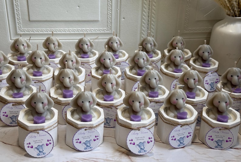 May include: A collection of small, white, hexagonal boxes with a gray elephant candle inside each. The boxes have a brown twine bow and a small tag that says "Baby Shower" with a gray elephant graphic.