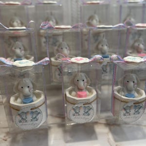 May include: A collection of small, clear boxes with white lids, each containing a small, white elephant figurine holding a blue or pink heart. The boxes are decorated with a pink and blue ribbon and a label that says "Laura & Ayden".