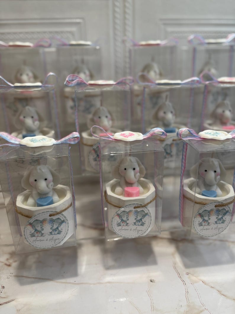 May include: A collection of small, clear boxes with white lids, each containing a small, white elephant figurine holding a blue or pink heart. The boxes are decorated with a pink and blue ribbon and a label that says "Laura & Ayden".