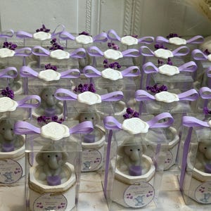 May include: A collection of small, clear boxes with purple ribbon tied around them. Each box contains a small, gray elephant figurine holding a purple heart. The boxes are decorated with a white wax seal and a small, white tag with the text "Baby Shower" printed on it.