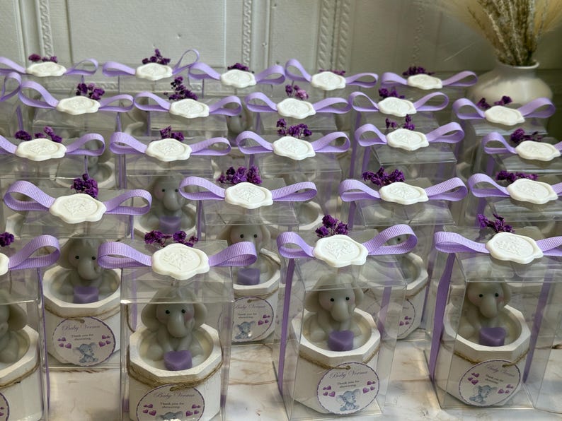 May include: A collection of small, clear boxes with purple ribbon tied around them. Each box contains a small, gray elephant figurine holding a purple heart. The boxes are decorated with a white wax seal and a small, white tag with the text "Baby Shower" printed on it.