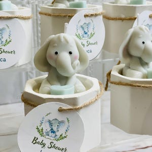 May include: White ceramic candle holders with a small elephant figurine on top. Each holder has a brown twine accent and a round tag with a blue and green elephant illustration and the text "Baby Shower Luca".