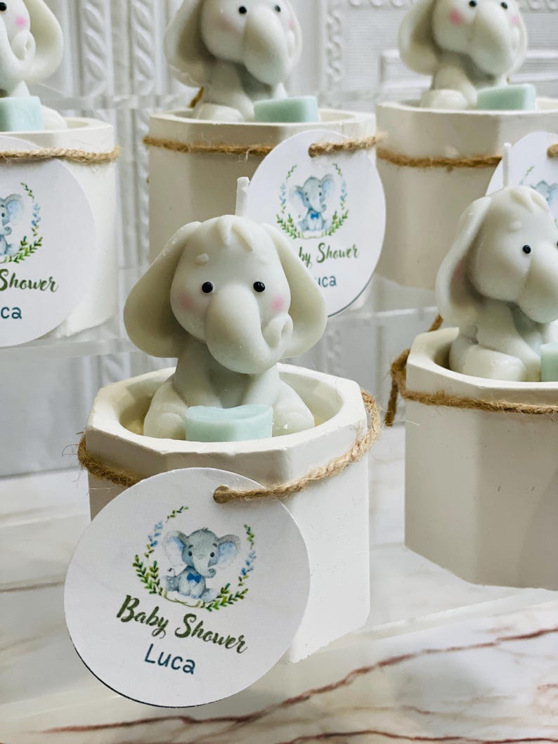 May include: White ceramic candle holders with a small elephant figurine on top. Each holder has a brown twine accent and a round tag with a blue and green elephant illustration and the text "Baby Shower Luca".