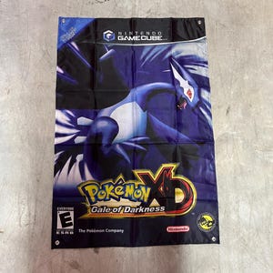 May include: A rectangular Pokémon XD: Gale of Darkness banner. The banner features a dark blue and purple background with a stylized image of a Pokémon. The Nintendo GameCube logo is at the top.