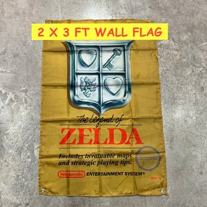 May include: A 2 x 3 foot wall flag featuring the Legend of Zelda logo. The logo is a silver shield with a key, a heart, and a lion. The flag is gold with the text "The Legend of Zelda" in black and the Nintendo Entertainment System logo.