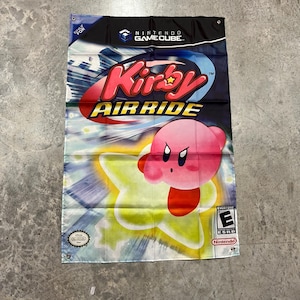 May include: A rectangular wall banner featuring the Kirby Air Ride video game artwork. The design displays the pink Kirby character riding a yellow star, with the Nintendo GameCube logo at the top and an ESRB E rating symbol at the bottom right.