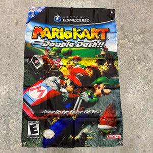 May include: A black and red Nintendo GameCube Mario Kart Double Dash!! banner with a colorful image of Mario, Luigi, and other characters racing on a track. The banner has the text "Team Up for Twice the Fun!" and an ESRB rating of "Everyone".