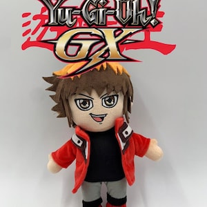 May include: A Yu-Gi-Oh! GX character plush doll, dressed in a red jacket, black shirt, and grey trousers. The doll has brown hair and red shoes. The Yu-Gi-Oh! GX logo is in the background.