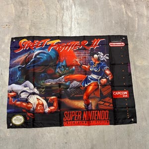 Könnte beinhalten: Schwarzes Banner mit dem Street Fighter II Super Nintendo-Spiel. Das Banner zeigt Charaktere in Aktion, mit dem Spieltitel in Rot und Weiß. Die Logos von Nintendo und Capcom sind sichtbar.