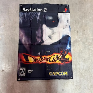 May include: A black banner featuring the PlayStation 2 logo and the title "Devil May Cry 2." The banner displays a character's face with white hair and a red eye. The Capcom logo is visible.