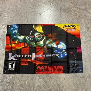 Puede incluir: Una bandera negra y roja con un personaje verde y azul en el centro. La bandera dice "Killer Instinct" y "Super Nintendo Entertainment System".