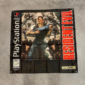 Op de afbeelding: Een zwart-rode Resident Evil videogamevlag met het PlayStation-logo, een leeftijdsclassificatie voor volwassenen en het Capcom-logo. De vlag toont een personage dat een geweer vasthoudt voor een zombie-achtige achtergrond.