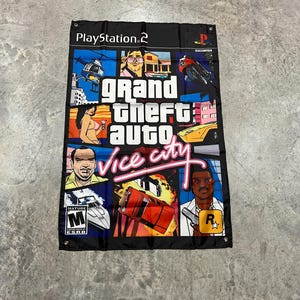 May include: A black and colourful flag featuring the text "Grand Theft Auto: Vice City" and the PlayStation 2 logo. The flag depicts various characters and scenes from the video game, including a helicopter, a car, and a man with a gun.