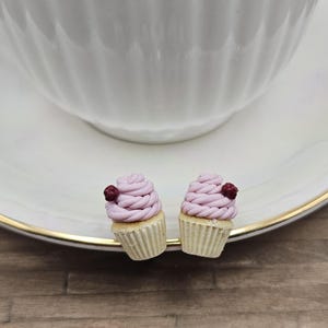 May include: Two miniature cupcakes with light pink frosting and a dark red berry on top. The cupcakes sit on a white plate with a gold rim, near a white teacup. The cupcakes have a beige paper cup base.