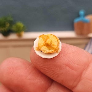 May include: Miniature pancake with syrup on a small white plate. The tiny breakfast food is held between fingers, showcasing the detail of the miniature food item. The pancake is golden brown with syrup drizzled on top, a charming addition to a dollhouse.