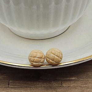 May include: Two small, round, light-colored objects with a crosshatch pattern sit on a white saucer with a gold rim. A white teacup is partially visible in the background. The objects appear to be decorative or edible.