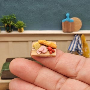 May include: Miniature charcuterie board with a slice of cheese, salami, baguette, and tomatoes. The wooden board is held in a hand, showcasing the tiny food items. The background features a blue wall and small decorative items.