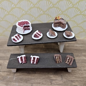 May include: Miniature cakes and slices of cake are displayed on a two-tiered dark gray table. The cakes are white and chocolate with red and brown fillings. The table has white legs and sits on a wooden surface.