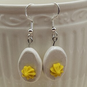 May include: A pair of handmade earrings featuring a deviled egg design. Each earring has a white, oval-shaped egg with a yellow yolk detail. The earrings hang from silver-colored fish hook ear wires.