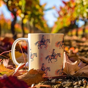 Puede incluir: Una taza de cer&aacute;mica blanca con asa, con un patr&oacute;n repetido de vaqueros montando a caballo. El fondo es de color beige claro con manchas marrones. La taza est&aacute; sentada sobre una cama de hojas de oto&ntilde;o.