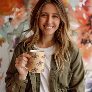 Puede incluir: Una taza de cer&aacute;mica blanca con un dise&ntilde;o marr&oacute;n y amarillo que presenta un patr&oacute;n repetido de una persona montando a caballo. La taza est&aacute; siendo sostenida por una persona con cabello largo y casta&ntilde;o.