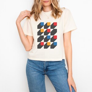 May include: Cream-colored t-shirt featuring a geometric design of alternating black, red, yellow, and blue squares. The squares have a 3D effect, creating a visually striking pattern. The shirt is paired with blue jeans.