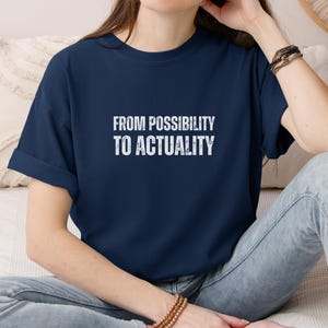 May include: A navy blue short-sleeved t-shirt featuring the white text "FROM POSSIBILITY TO ACTUALITY" printed in a distressed, weathered font across the center of the chest. The shirt has a crew neckline and a relaxed fit with rolled sleeves.