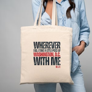 May include: A beige tote bag with the text "WHEREVER I GO, I TAKE A LITTLE PIECE OF WASHINGTON, D.C. WITH ME" in black and red. The bag has two handles and is held by a person.