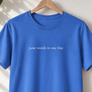 May include: A bright blue short-sleeved t-shirt featuring the white text "your words in one line" printed in a simple, lowercase serif font across the centre of the chest. The garment has a classic crew neckline and is displayed on a wooden hanger.