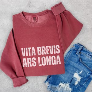 May include: A maroon sweatshirt with the text "VITA BREVIS ARS LONGA" in white, paired with denim shorts. The sweatshirt has a crew neck and long sleeves with folded cuffs. The shorts are blue denim with distressed details.