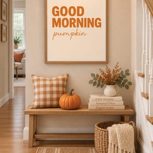 May include: A framed wall art print featuring the orange text GOOD MORNING pumpkin, displayed above a wooden bench with a plaid cushion, a pumpkin, a white vase with dried foliage, and a stack of books labelled Cozy Seasons, Autumn Vibes, Happy Home.