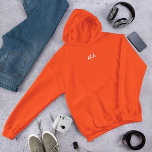 Puede incluir: Sudadera con capucha de color naranja brillante con la palabra "bitch" en escritura blanca. La sudadera con capucha tiene un bolsillo delantero y una capucha. La imagen también muestra vaqueros azules, zapatillas grises, un cinturón negro, auriculares, un smartphone y una cámara.