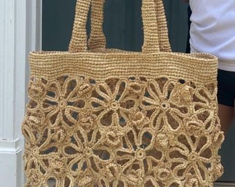 May include: A beige woven tote bag with a floral design. The bag has two handles and a large, open compartment. The floral pattern is made of woven material, creating a three-dimensional effect. The bag is made of a natural fiber material.