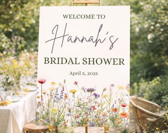 May include: A welcome sign for Hannah's bridal shower, displayed on a gold easel. The sign features the text "Welcome to Hannah's Bridal Shower" and the date April 5, 2025. A floral border with colorful wildflowers decorates the bottom.