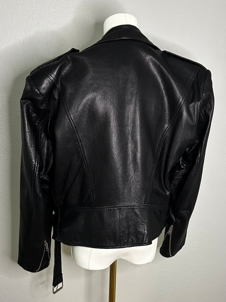 May include: Black leather jacket with a belted waist and zippered pockets. The jacket has a slightly oversized fit and features exaggerated shoulder pads.